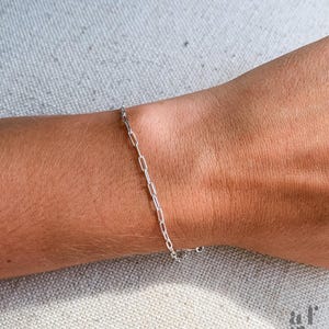 Op de afbeelding: Een delicate zilveren kettingarmband met rechthoekige schakels wordt getoond op een pols. De armband is eenvoudig en elegant, met een kleine sluiting. De achtergrond is een neutrale, getextureerde stof.