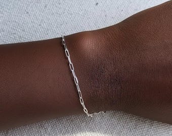 May include: A delicate silver chain bracelet with an elongated oval link design. The bracelet is shown on a dark skin tone wrist, with a simple, elegant aesthetic. The bracelet is a classic piece of jewelry.