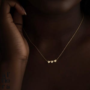 Può includere: Una delicata collana d'oro con tre ciondoli a forma di cuore, ognuno con un'iniziale. La collana è mostrata su uno sfondo scuro, evidenziando i dettagli raffinati e l'elegante design del gioiello. Le iniziali sono E, H e J.