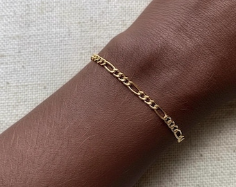 May include: A gold-colored Figaro chain bracelet. The bracelet is displayed on a brown arm. The chain links alternate between two different shapes, creating a classic and elegant design. The bracelet is a simple yet stylish accessory.