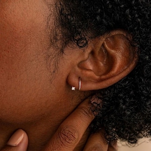 May include: A close-up of a person's ear with a small, silver hoop earring and a small, square-cut diamond stud earring. The person's skin is a warm brown tone, and their dark, curly hair is visible.
