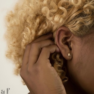 May include: A close-up of a person's ear with a small, gold-colored letter "M" stud earring. The person's hand is near the ear, and the background is a soft, neutral color. The hair is blonde and curly. The earring is a minimalist jewelry piece.