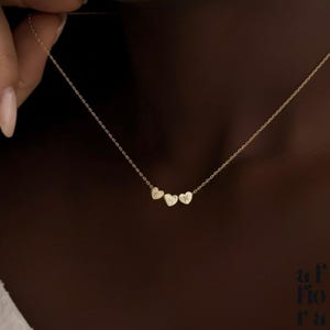 May include: A delicate gold necklace with three heart-shaped pendants. Each heart has an initial engraved on it. The necklace is shown against a dark background, highlighting the jewellery's fine details.