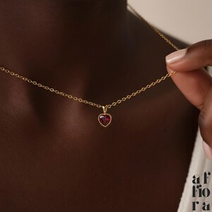 May include: A delicate gold necklace featuring a heart-shaped pendant with a red gemstone center. The pendant is surrounded by a halo of small, sparkling accents. The necklace is held by a hand, showcasing the fine chain and pendant design.