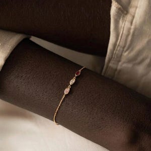 May include: A delicate gold bracelet featuring three colorful gemstones: a pink, a clear, and a red stone. The bracelet is shown on a dark skin tone, with a neutral-colored shirt in the background. The gemstones are oval-shaped.