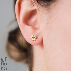 May include: Gold-toned paw print earrings with the letter 'A' engraved in the center. The earrings are shown on a person's earlobe.