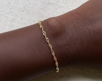May include: A delicate gold-colored bracelet with a twisted rope design. The bracelet is shown on a dark brown skin tone. The jewelry piece is simple and elegant, suitable for everyday wear or special occasions.