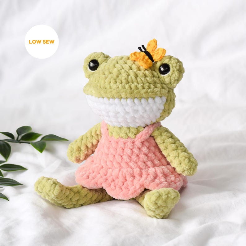 A handmade crochet frog amigurumi in pink dress with yellow butterfly on its head, made in light green chenille yarn, low sew advanced beginner friendly frog crochet pattern