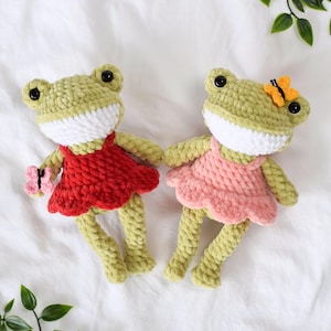 Two cute handmade crochet frog amigurumi plush toys in red and pink dresses, with butterfly on their heads. Both frogs have green bodies, white mouths, and black eyes.