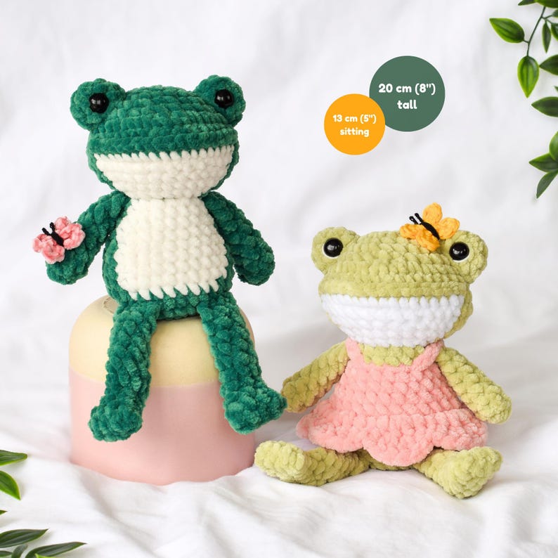 Two handmade crochet frog plushies. One is green and white, 8 inches tall, with a pink butterfly. The other is light green with a pink dress and a yellow butterfly on its head. Sitting height is 5 inches. 2 in 1 low sew crochet pattern PDF download.