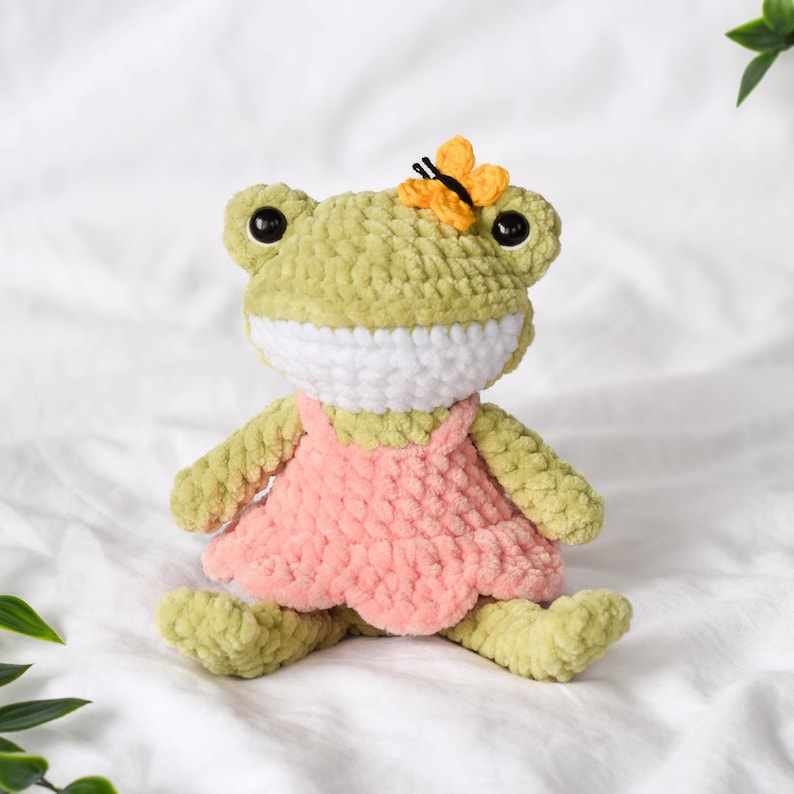 A handmade crochet frog doll in shades of green and pink. The frog has large black eyes, a white mouth, and a small yellow butterfly on its head. It is wearing a pink dress. Cottagecore animal plush toy crochet pattern.