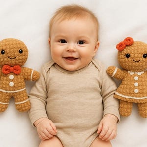 May include: A smiling baby wearing a beige onesie lies between two crocheted gingerbread dolls. The dolls are brown with white accents, and one has a red bow tie, the other a red bow.
