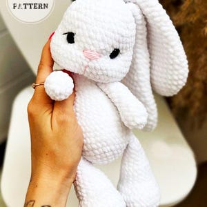 Peut inclure: Peluche lapin blanc en crochet avec des yeux noirs et un nez rose. Le lapin est tenu par une main.
