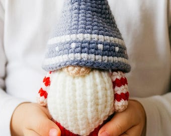 Op de afbeelding: Een handgemaakte gehaakte kabouterpop met een blauw-witte hoed, een crèmekleurige baard en een rode body met witte accenten. De pop wordt in de handen van een kind gehouden.