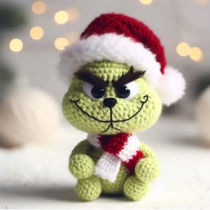 Puede incluir: Un personaje verde tejido a ganchillo con un gorro de Papá Noel rojo y blanco y una bufanda roja y blanca. El personaje tiene ojos negros, una sonrisa cosida y cejas marrones. El fondo está borroso con luces suaves y desenfocadas.