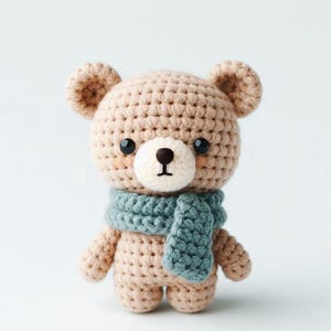 Puede incluir: Un pequeño oso de crochet marrón que lleva una bufanda azul. El oso tiene una cara blanca con ojos negros y una nariz negra.
