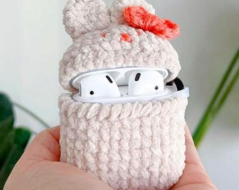 Pode incluir: Uma capa de Airpods de crochê branco com um design de coelho rosa e branco. A capa está aberta e os Airpods brancos são visíveis no interior.