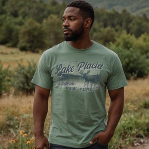 May include: A sage green short-sleeve t-shirt featuring a graphic print of a moose standing in a lake. The text reads "MOOSE COUNTRY LAKE PLACID NEW YORK" in a stylized font. The design includes horizontal lines and a nature-inspired scene.