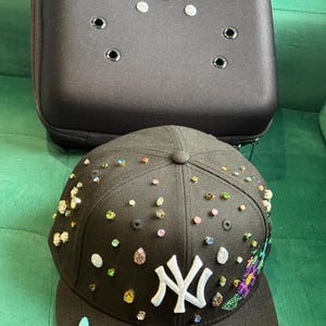 Puede incluir: Gorra de béisbol negra con el logo "NY" blanco y adornos coloridos. La gorra tiene una visera plana y está adornada con varias piedras preciosas y bordados florales. Un estuche negro con agujeros decorativos está en el fondo.