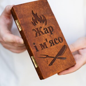 May include: A rectangular brown wooden box with two gold-colored metal hinges on the side. The lid features an engraved flame icon, the text "Жар і м'ясо", and a crossed carving fork and knife graphic. The wood has a visible grain and stained finish.