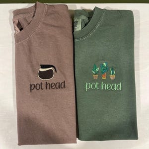 May include: Two t-shirts with the text "pot head" embroidered on them. One shirt is brown with a coffee pot design and the other is green with a potted plant design.