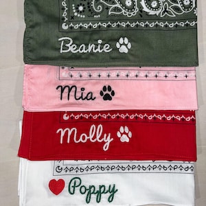 May include: A stack of four folded fabric bandanas in olive green, light pink, red, and white. Each bandana features a paisley border and is embroidered with a name and a symbol: Beanie with a paw, Mia with a paw, Molly with a paw, and Poppy with a heart.