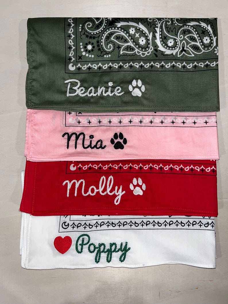 May include: A stack of four folded fabric bandanas in olive green, light pink, red, and white. Each bandana features a paisley border and is embroidered with a name and a symbol: Beanie with a paw, Mia with a paw, Molly with a paw, and Poppy with a heart.