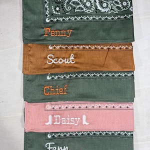 May include: A stack of five folded fabric bandanas in olive green, brown, and dusty pink. Each bandana features a printed paisley or geometric border and is embroidered with a name: Penny, Scout, Chief, Daisy, or Fern in orange or white thread.