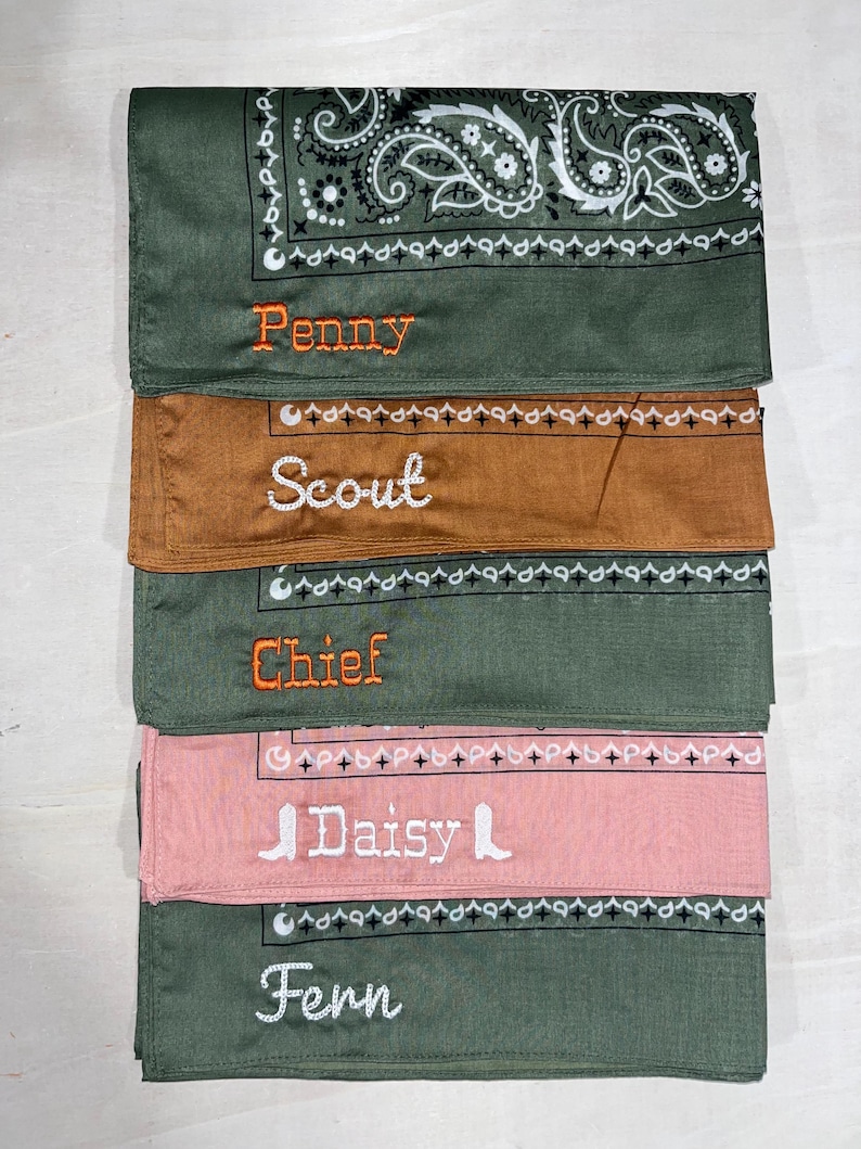 May include: A stack of five folded fabric bandanas in olive green, brown, and dusty pink. Each bandana features a printed paisley or geometric border and is embroidered with a name: Penny, Scout, Chief, Daisy, or Fern in orange or white thread.