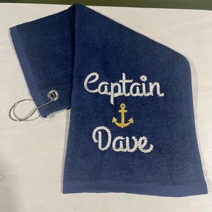 May include: Navy blue golf towel with the embroidered text "Captain Dave" in white script. A gold anchor design is between the words. The towel has a metal grommet and a silver clip.
