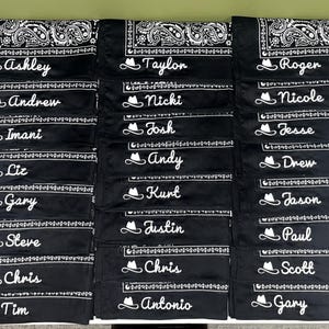 May include: A collection of black bandanas featuring a white paisley border pattern. Each bandana is personalized with a white embroidered cowboy hat icon followed by a unique name in a cursive script font, including Ashley, Taylor, Roger, Andrew, Nicki, Nicole, Imani, Josh, Jesse, Liz, Andy, Drew, Gary, Kurt, Jason, Steve, Justin, Paul, Chris, Chris, Antonio, Tim, and Scott.