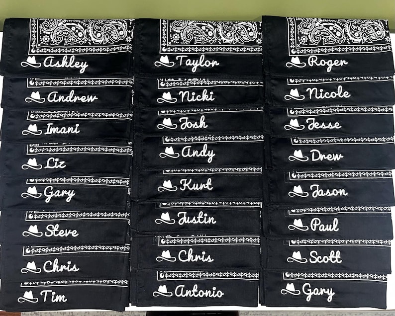 May include: A collection of black bandanas featuring a white paisley border pattern. Each bandana is personalized with a white embroidered cowboy hat icon followed by a unique name in a cursive script font, including Ashley, Taylor, Roger, Andrew, Nicki, Nicole, Imani, Josh, Jesse, Liz, Andy, Drew, Gary, Kurt, Jason, Steve, Justin, Paul, Chris, Chris, Antonio, Tim, and Scott.
