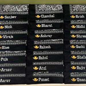 May include: A collection of black bandanas featuring a white paisley pattern along the edges. Each bandana is personalized with an embroidered yellow cowboy hat icon followed by a unique name in white text, including Sanjeev, Chanchal, Krish, Nick, Bharat, Nidhi, Viraja, Ashray, Shreeda, Rhea, Rakesh, Sachi, Puja, Rahul, Shelley, Arnay, Atul, Rajiv, Aaray, Puneet, and Umang.