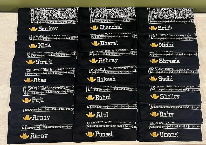 May include: A collection of black bandanas featuring a white paisley pattern along the edges. Each bandana is personalized with an embroidered yellow cowboy hat icon followed by a unique name in white text, including Sanjeev, Chanchal, Krish, Nick, Bharat, Nidhi, Viraja, Ashray, Shreeda, Rhea, Rakesh, Sachi, Puja, Rahul, Shelley, Arnay, Atul, Rajiv, Aaray, Puneet, and Umang.