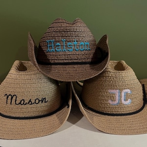 May include: Three straw cowboy hats with personalized names embroidered on the front. The hats are in shades of brown and tan, with black bands and chin straps. One hat has "Halston" in turquoise, another "Mason" in black, and the third "JC" in pink and blue.