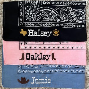 May include: Three folded bandanas in black, light pink, and light blue. The black bandana features a white paisley pattern, a gold embroidered Texas shape, the name Halsey, and a gold star. The pink one has black embroidery of boots and the name Oakley. The blue one has a brown cowboy hat and the name Jamie.