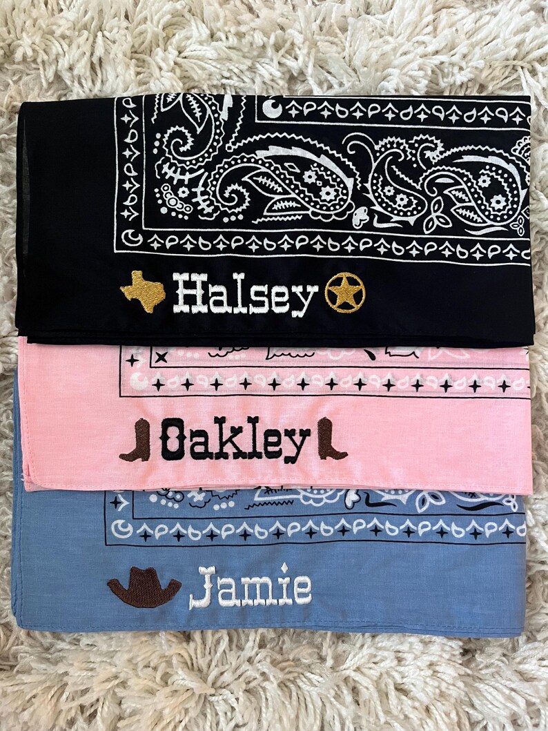 May include: Three folded bandanas in black, light pink, and light blue. The black bandana features a white paisley pattern, a gold embroidered Texas shape, the name Halsey, and a gold star. The pink one has black embroidery of boots and the name Oakley. The blue one has a brown cowboy hat and the name Jamie.