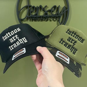 May include: Two trucker hats, one black and one olive green camouflage, are held up. Both hats have the phrase "tattoos are trashy" embroidered in white and black, respectively. The hats have a mesh back and a curved brim.
