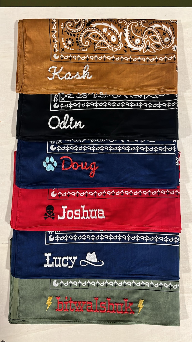 May include: A stack of six folded bandanas in brown, black, navy, red, blue, and olive green. Each features a white paisley border and embroidered names: Kash, Odin, Doug with a paw print, Joshua with a skull, Lucy with a hat, and bitwalshuk with lightning bolts.