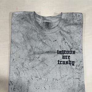 May include: A gray Comfort Colors crew neck t-shirt with a distressed, textured pattern. The shirt features the phrase "tattoos are trashy" in black gothic lettering. The shirt is folded neatly on a light wood surface.