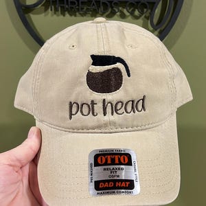 May include: Tan dad hat with the words "pot head" embroidered in brown, along with a coffee pot graphic. The hat has a curved brim and a tag that reads "OTTO" and "DAD HAT".