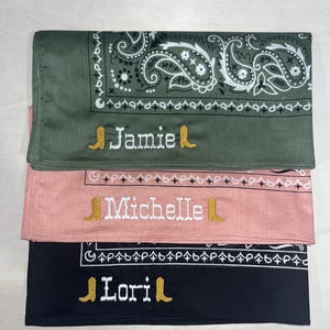 May include: Three folded bandanas in olive green, dusty pink, and black. Each features a white paisley border and is embroidered with a name in white thread, flanked by two small gold-colored cowboy boot icons. The names are Jamie, Michelle, and Lori.