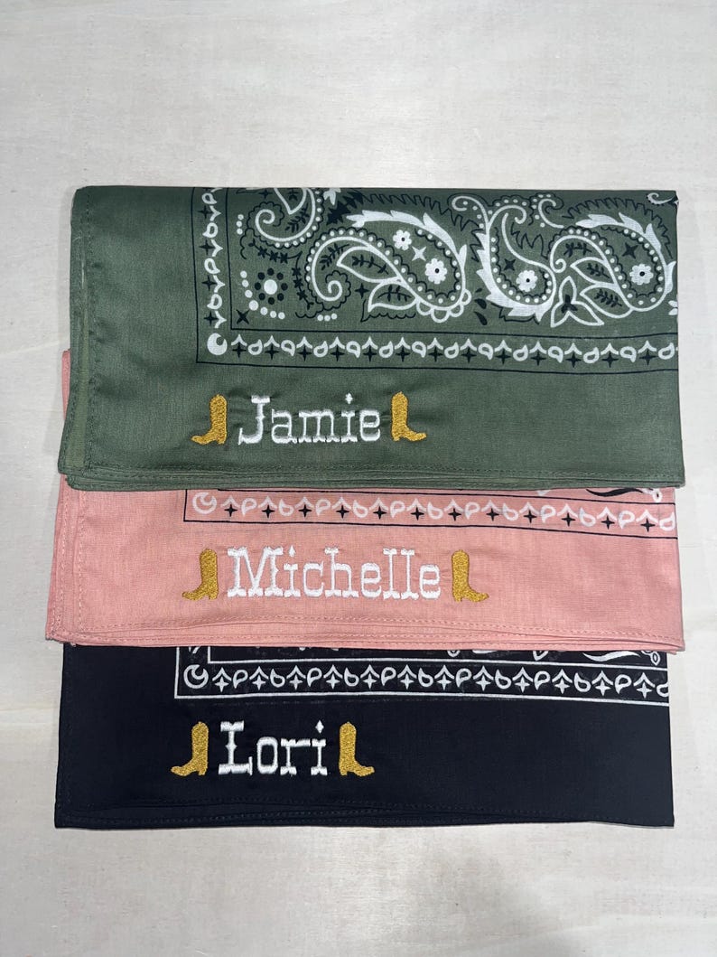May include: Three folded bandanas in olive green, dusty pink, and black. Each features a white paisley border and is embroidered with a name in white thread, flanked by two small gold-colored cowboy boot icons. The names are Jamie, Michelle, and Lori.