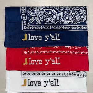 May include: Three folded bandanas in navy blue, red, and white, each featuring a traditional paisley border pattern. Embroidered on each is a gold cowboy boot icon followed by the text "love y'all" in a western-style font with contrasting thread.