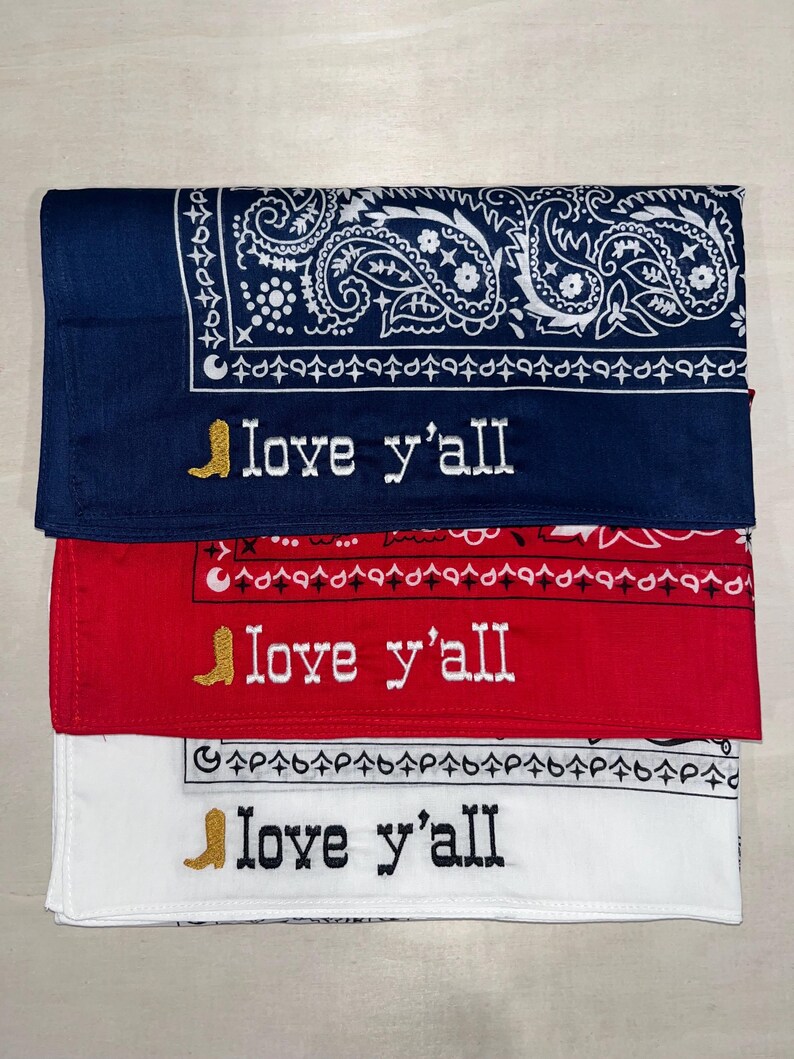 May include: Three folded bandanas in navy blue, red, and white, each featuring a traditional paisley border pattern. Embroidered on each is a gold cowboy boot icon followed by the text "love y'all" in a western-style font with contrasting thread.