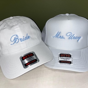 May include: Two white baseball caps. One cap has the word "Bride" embroidered in light blue script. The other cap is a trucker hat with "Mrs. Uray" embroidered in light blue script. Both caps have the "OTTO" brand label.