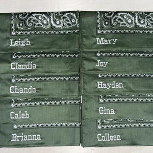 May include: Ten olive green bandanas arranged in two columns, each featuring a white paisley border pattern and a unique name embroidered in white thread. The names include Leigh, Mary, Claudia, Joy, Chanda, Hayden, Caleb, Gina, Brianna, and Colleen.