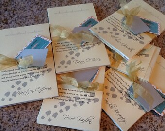 May include: A collection of cream-colored, personalized cards with gold ribbon and tags. Each card features a printed quote, footprints, and a name. The cards are likely for a special occasion, such as a memorial or celebration.