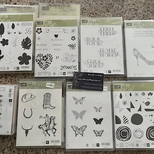 May include: A collection of ten clear plastic stamp sets with various designs. The designs include floral patterns, geometric shapes, hearts, butterflies, and a cowboy boot. The stamp sets are arranged in two rows on a light brown surface.