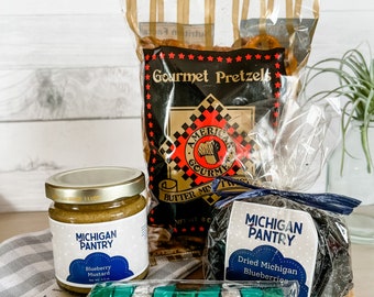 May include: A gift basket with Michigan Pantry products. The basket includes a jar of blueberry mustard, a bag of gourmet pretzels, a bag of dried Michigan blueberries, and a package of chocolate-covered blueberries.